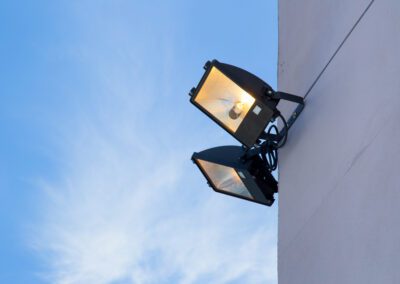 Two large industrial floodlights are mounted on a building exterior against a partly cloudy blue sky. One light is glowing, while the other remains off.